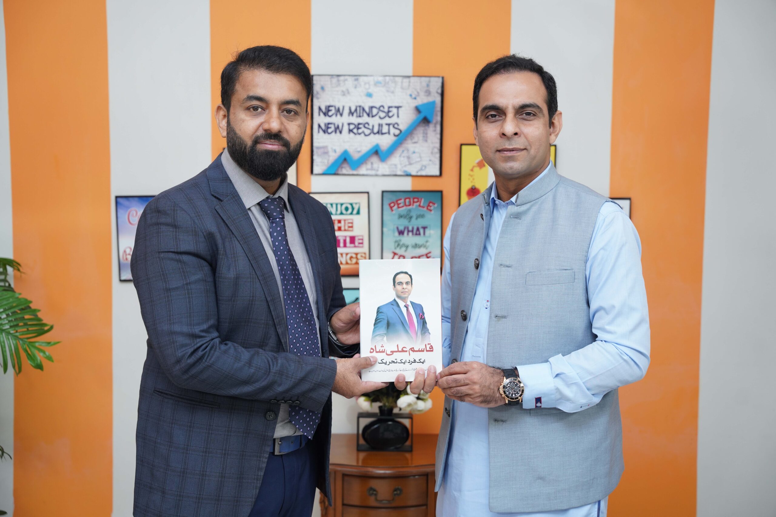 Munir Ahmad and Qasim Ali Shah holding a book together during a professional meeting, standing in front of motivational posters.
