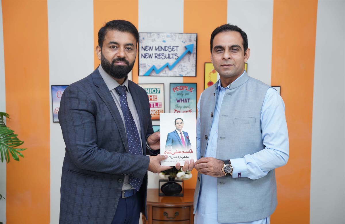 Munir Ahmad and Qasim Ali Shah holding a book together during a professional meeting, standing in front of motivational posters.