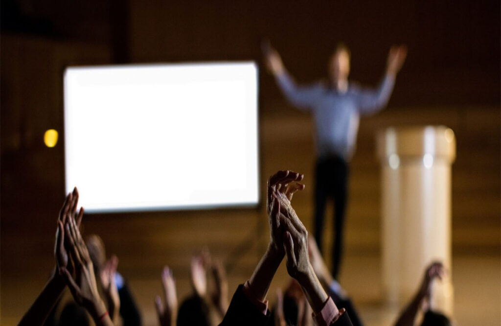 Audience applauding a Pakistani Motivational Speakers standing on stage beside a presentation screen.